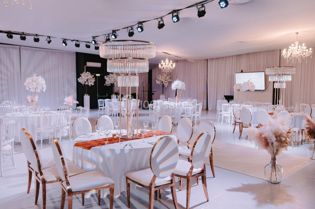 Salle de réception élégante avec tables rondes dressées, chaises blanches et décorations lumineuses et florales.