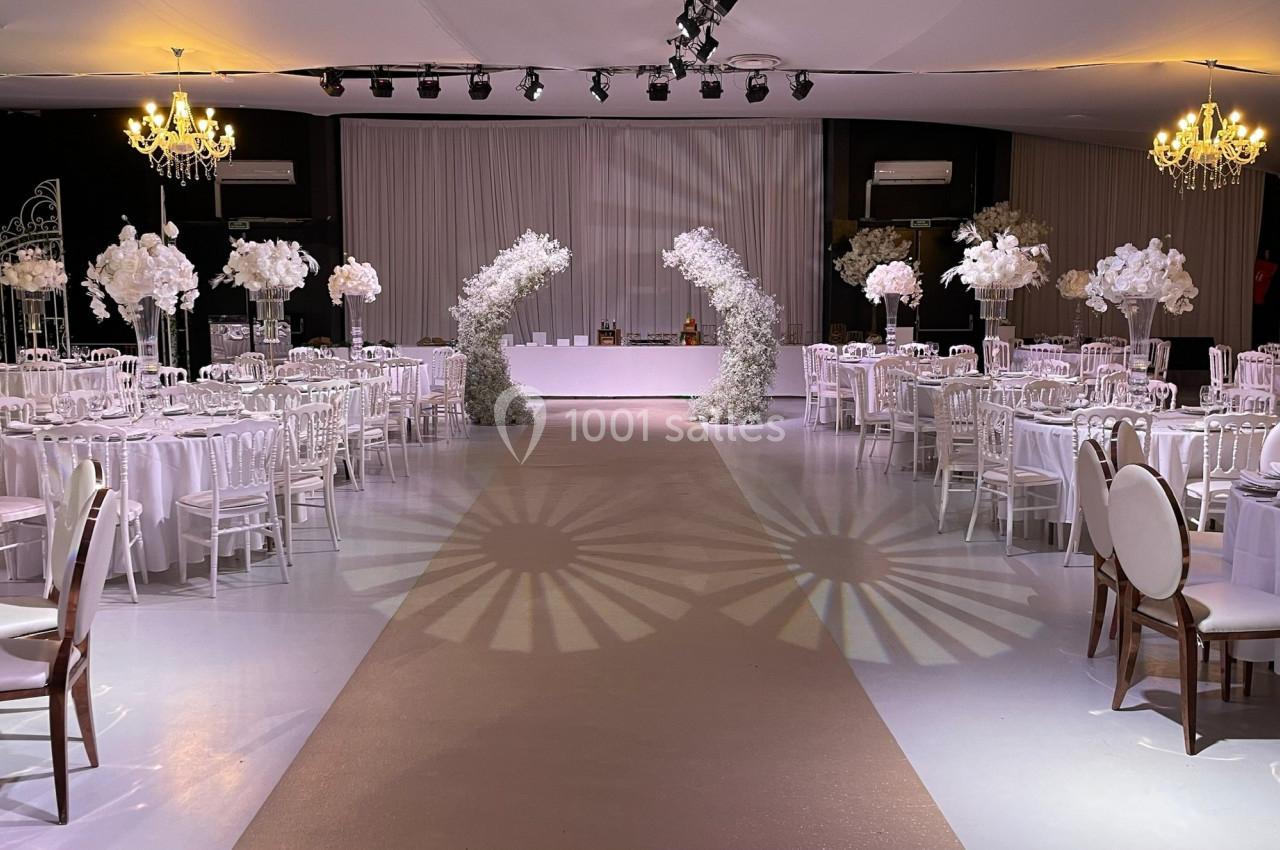 Salle de réception décorée avec des tables rondes, des chaises blanches et des arches florales blanches sous des lustres.