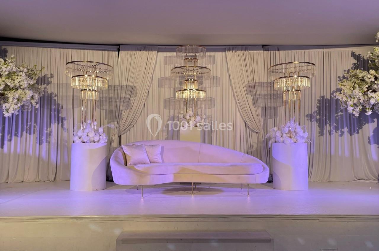 Décor élégant avec un canapé beige, des rideaux blancs, des lustres et des arrangements floraux blancs.