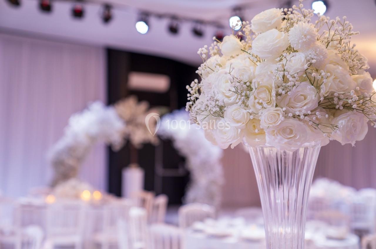 Arrangement floral blanc sur un vase transparent, avec en arrière-plan une arche décorée et des tables dressées.