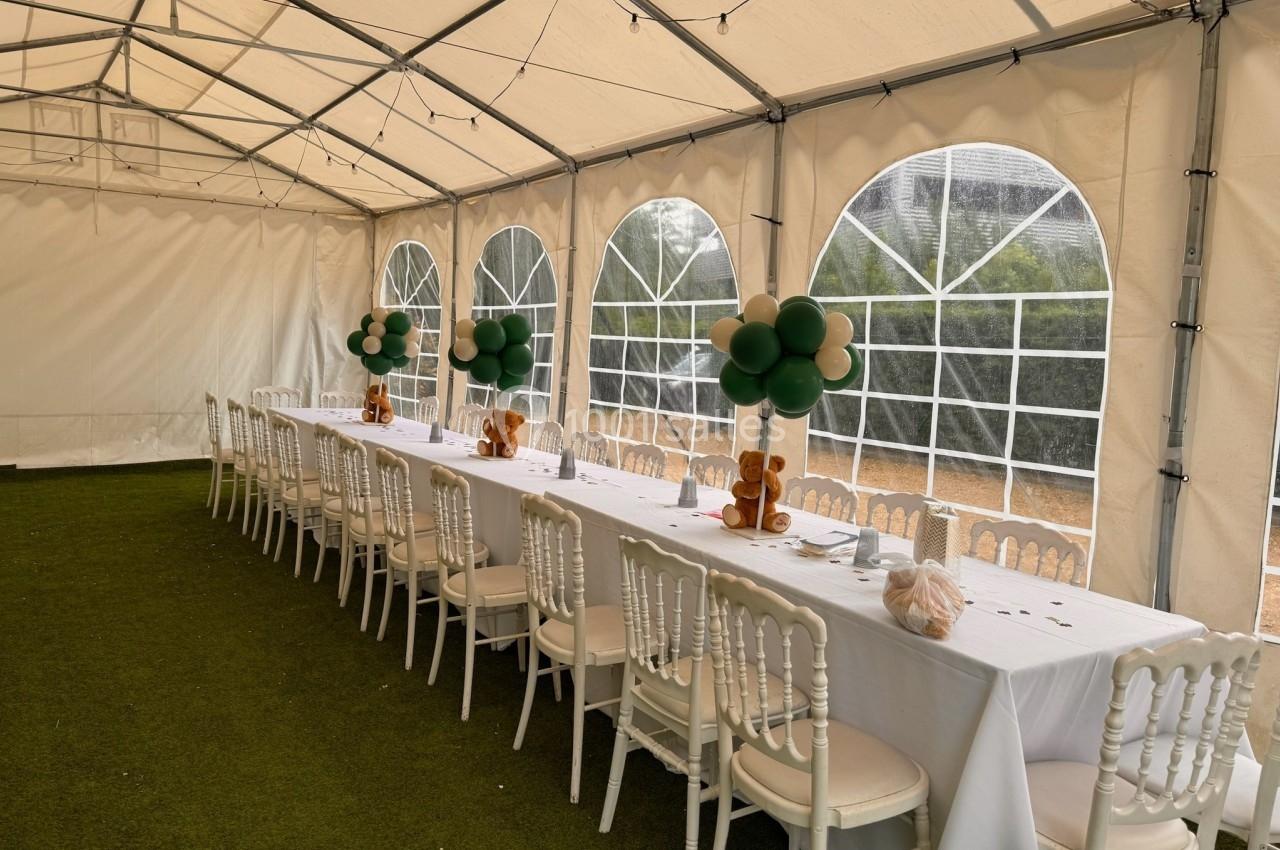 Grande table décorée avec nappes blanches, ballons et peluches, installée sous une tente avec sol en gazon artificiel.