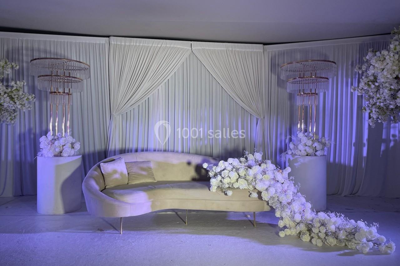 Canapé beige incurvé entouré de décorations florales blanches et de rideaux plissés dans un cadre élégant.