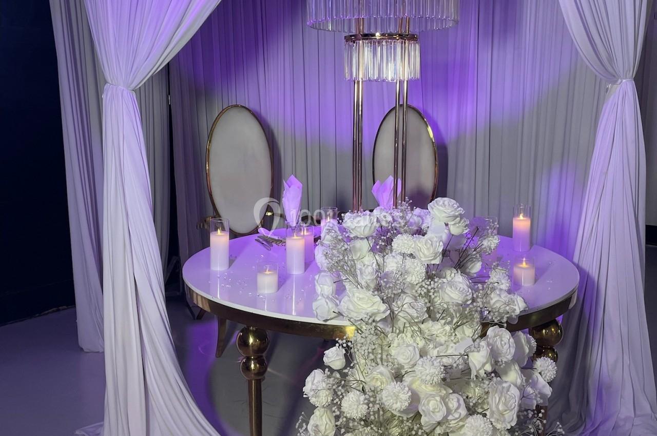 Décoration élégante avec table blanche, chaises ovales, fleurs blanches et rideaux drapés sous un éclairage violet.