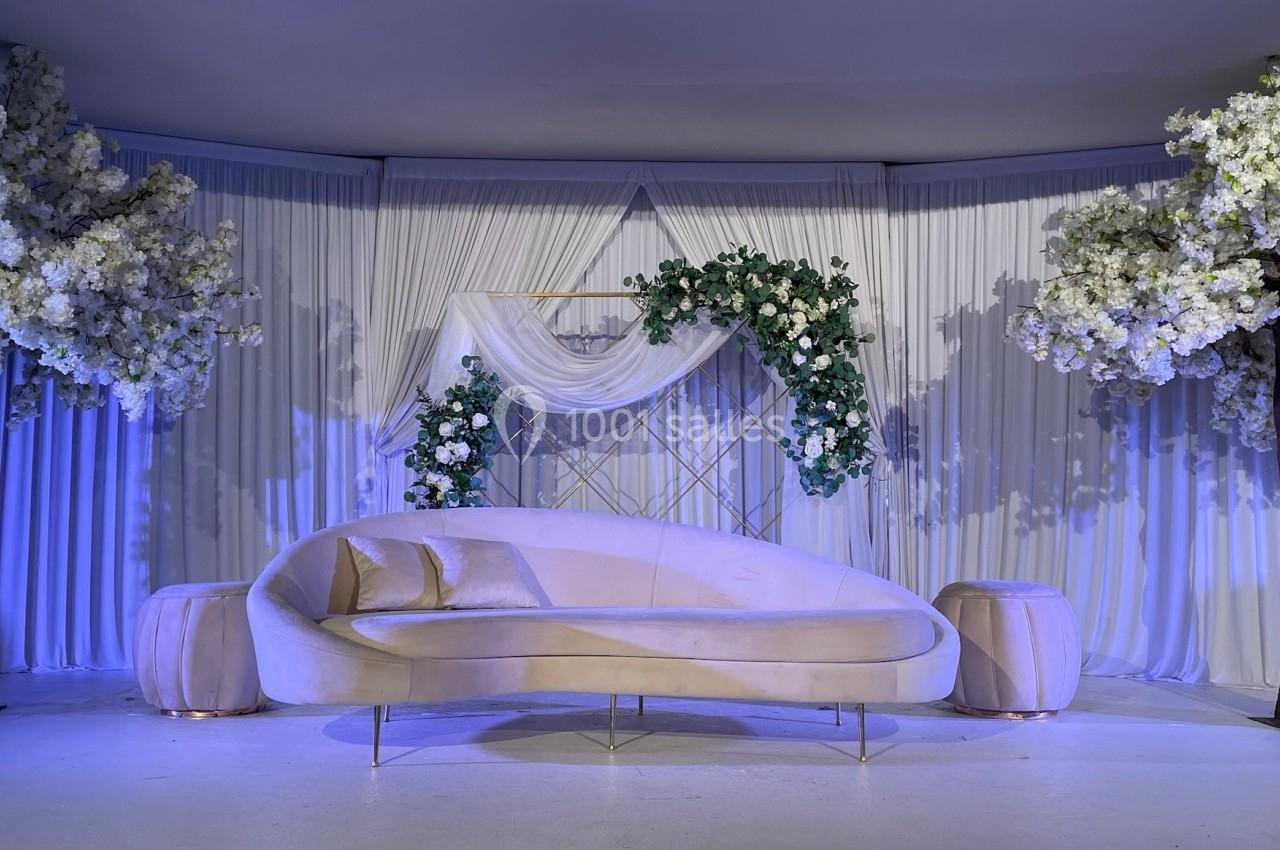 Canapé beige incurvé devant un décor élégant avec rideaux blancs, arches fleuries et arbres ornés de fleurs blanches.