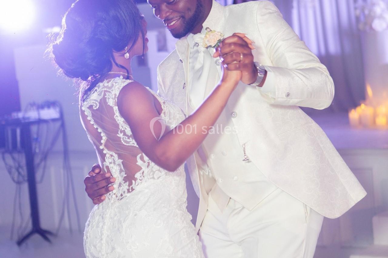 Un couple en tenue de mariage danse dans une salle décorée de fleurs blanches et éclairée par une lumière douce.