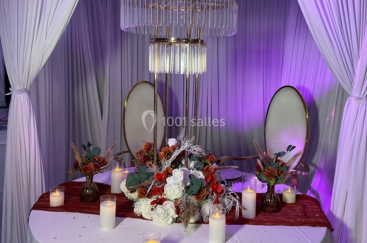 Table décorée avec des fleurs rouges et blanches, bougies allumées et lustre suspendu, entourée de rideaux blancs.