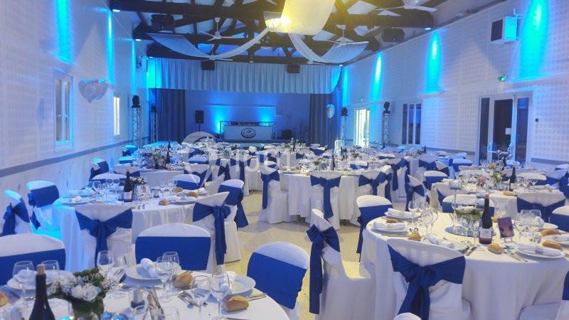 Salle de réception décorée avec des tables rondes, nappes blanches et nœuds bleus, éclairage tamisé bleu.