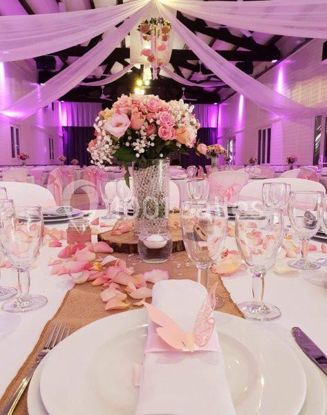 Salle de réception décorée dans des tons roses avec des fleurs, nappes blanches et éclairage tamisé.