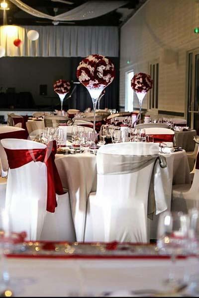Salle de réception décorée avec des tables rondes, nappes blanches, chaises ornées de rubans rouges et centres de table…