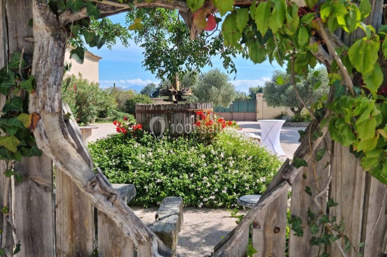 Vue d'un jardin fleuri à travers une ouverture dans une clôture en bois, avec une ancienne presse au centre.