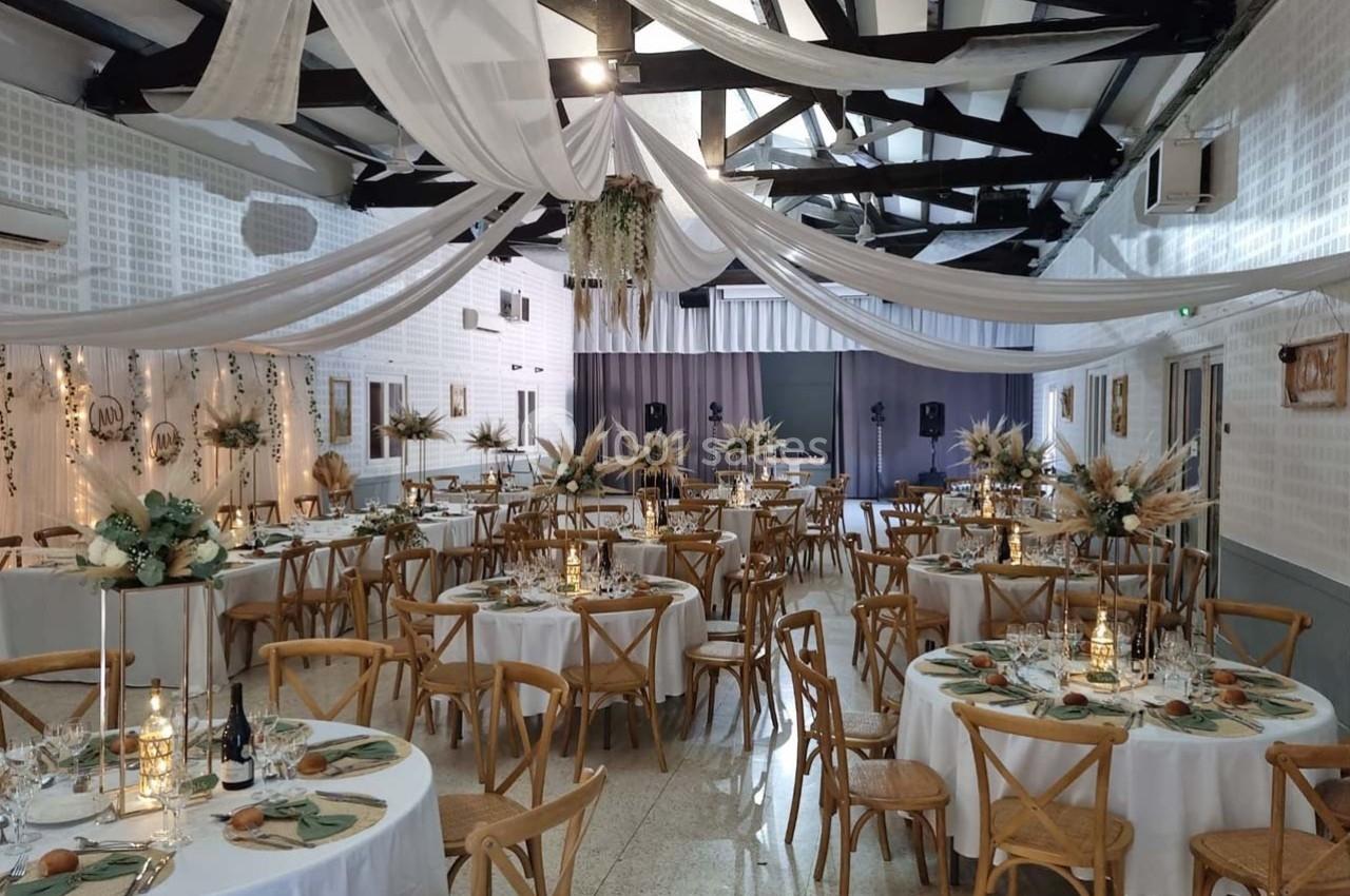 Salle de réception décorée pour un événement, avec tables rondes, chaises en bois et ornements floraux suspendus.