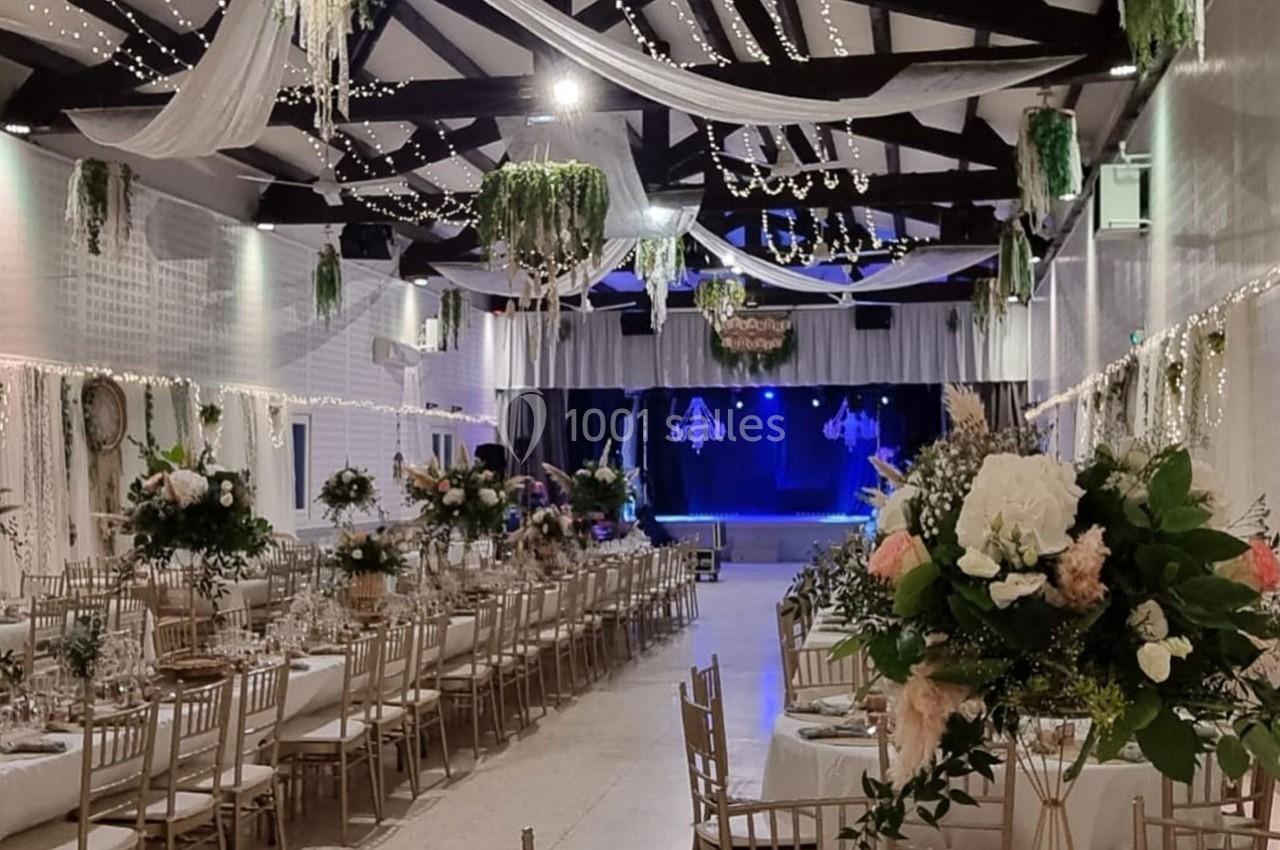 Salle de réception décorée avec des tables dressées, des fleurs, des guirlandes lumineuses et une scène en arrière-plan.
