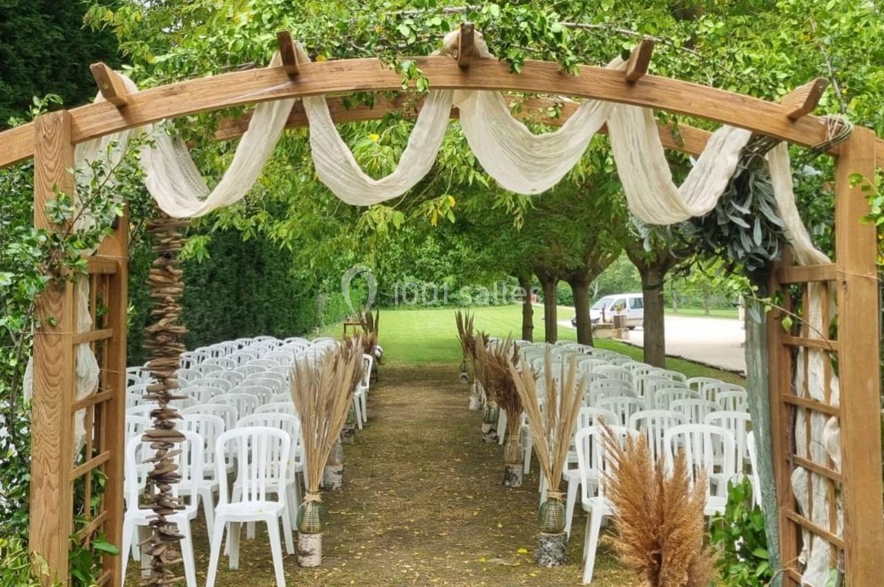 Allée décorée pour une cérémonie en plein air, avec arche en bois, voilages et rangées de chaises blanches.