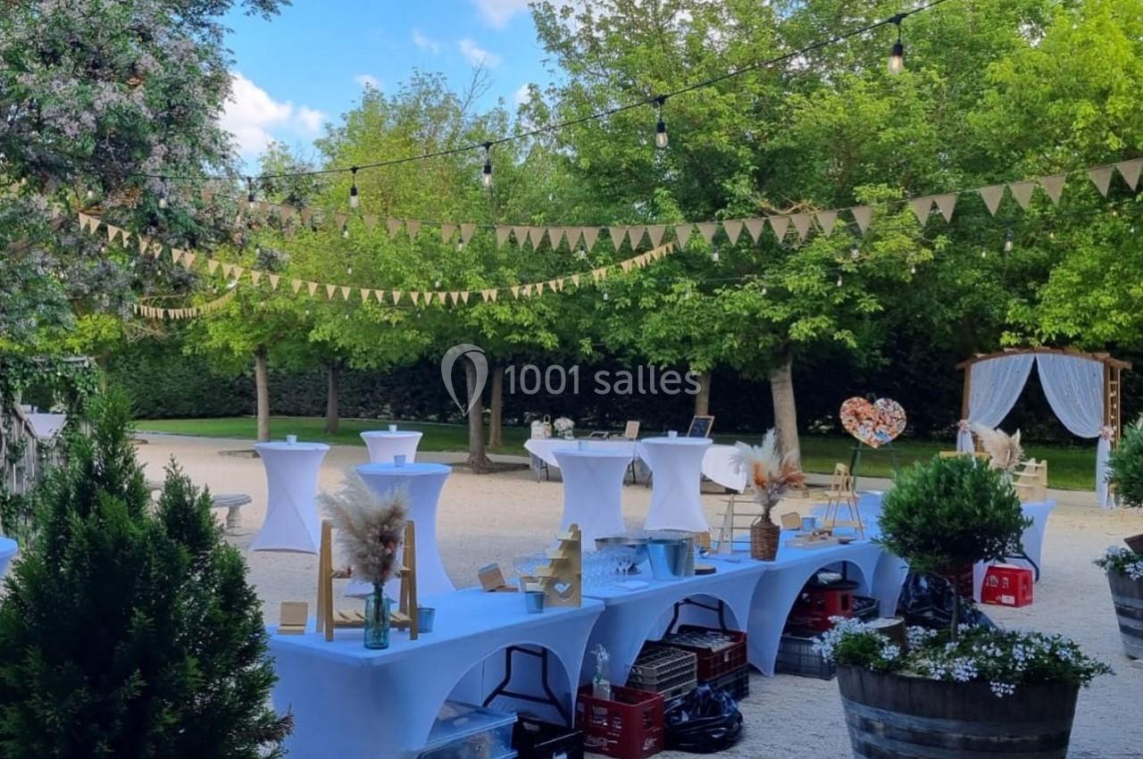 Décoration extérieure pour un événement avec tables hautes, guirlandes et verdure dans un jardin ensoleillé.