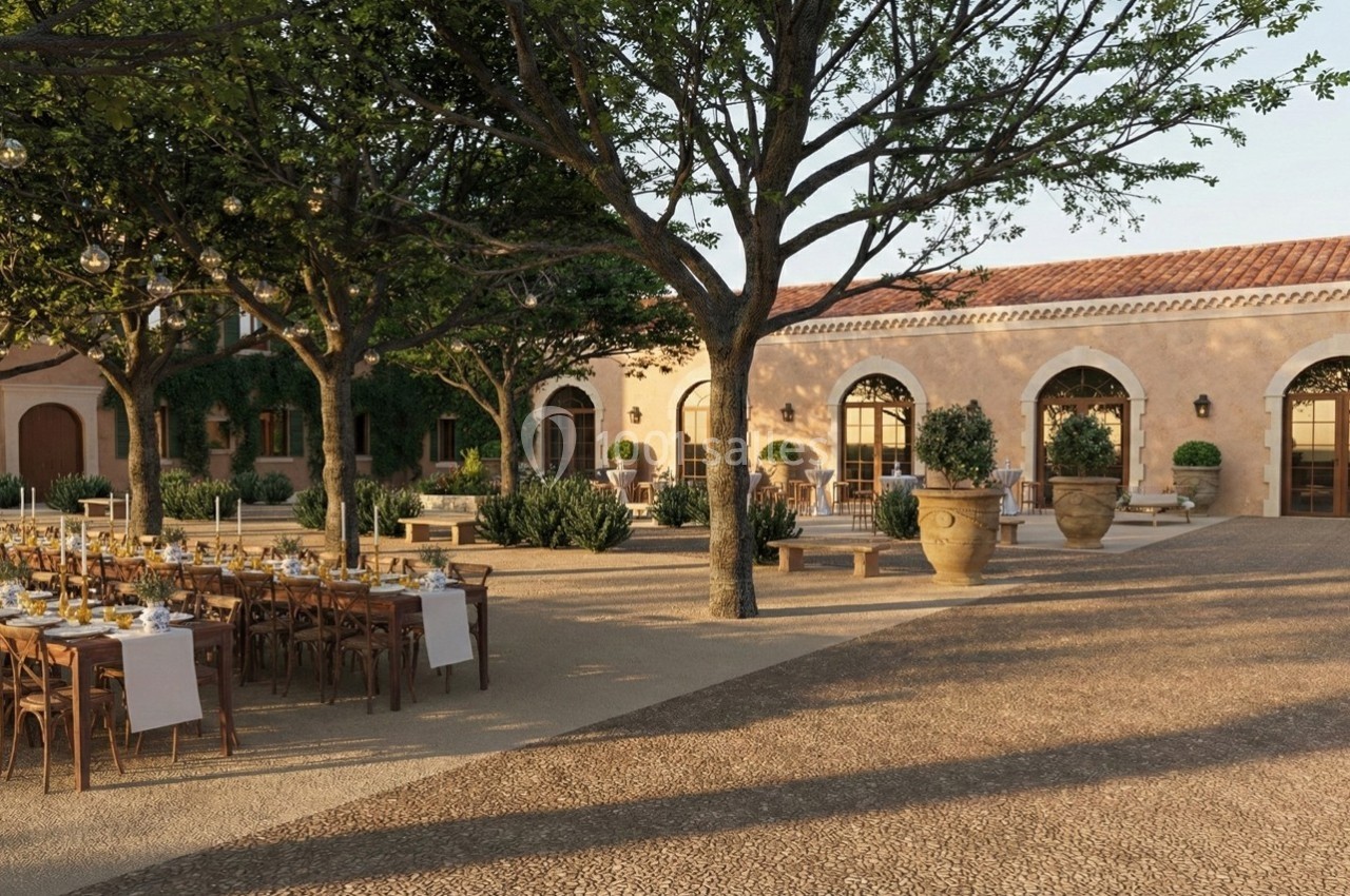 Cour extérieure aménagée avec tables dressées pour un événement, entourée d'arbres, de plantes et d'un bâtiment en pierre.