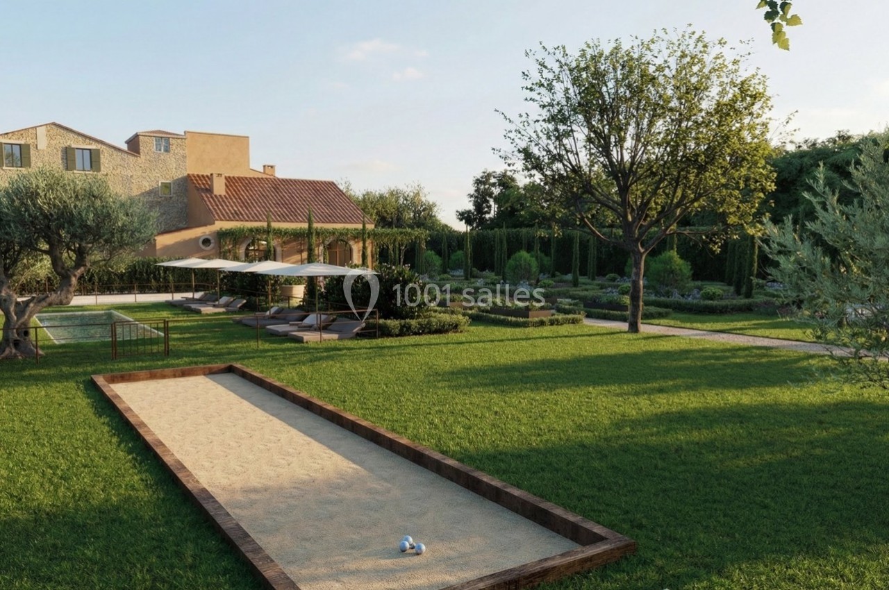 Jardin avec terrain de pétanque, pelouse, arbres, chaises longues sous parasols et maison en pierre en arrière-plan.