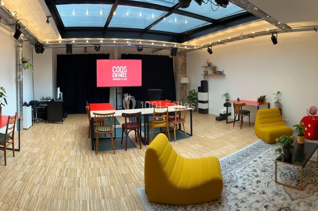 Salle lumineuse avec mobilier coloré, espace de travail collaboratif, scène et écran sous une verrière.