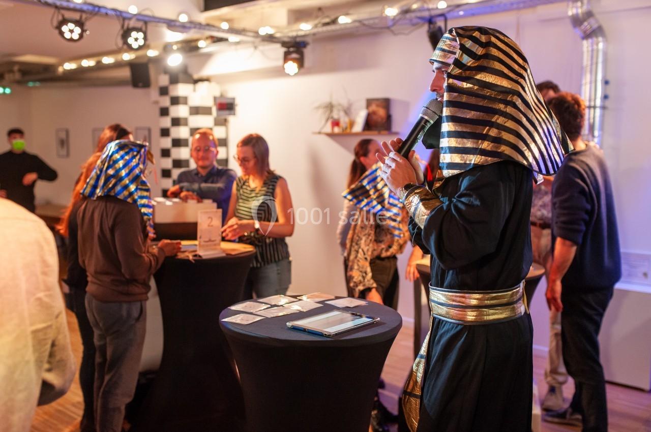 Un homme déguisé en pharaon anime une activité dans une salle avec des participants autour de tables hautes.