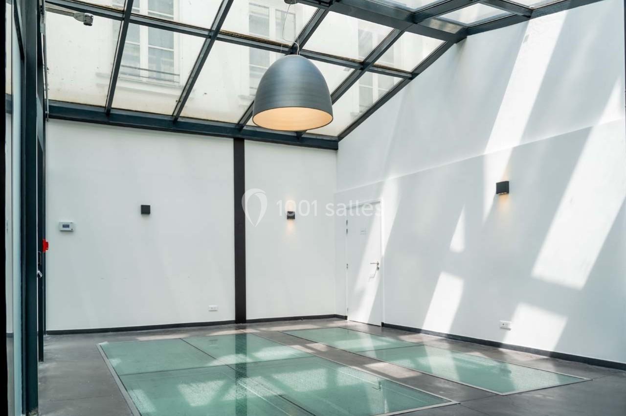 Salle lumineuse avec verrière, murs blancs, sol en verre et béton, éclairée par une suspension et des appliques murales.