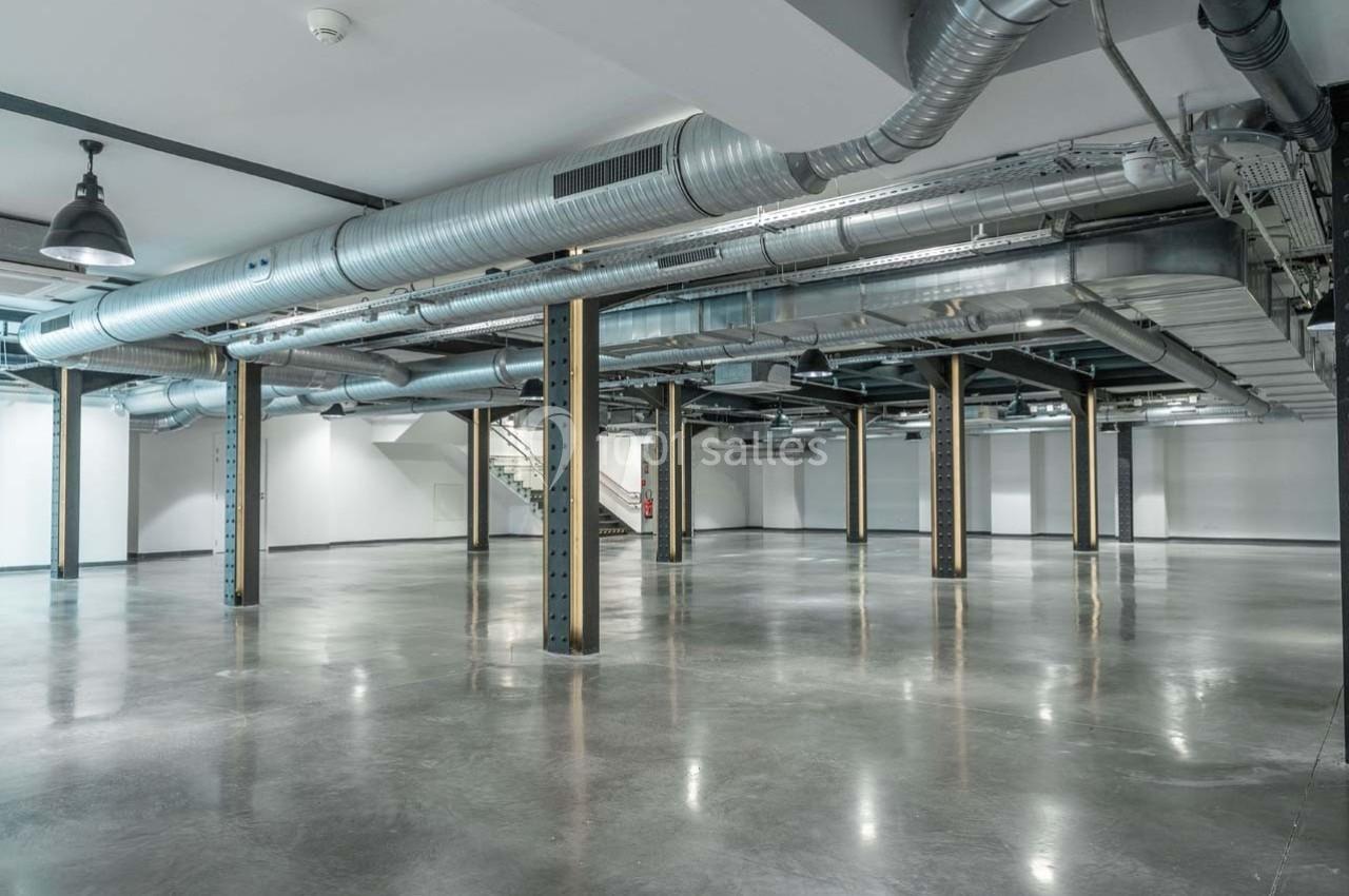 Intérieur d'un espace industriel vide avec sol en béton poli, colonnes métalliques et conduits apparents au plafond.