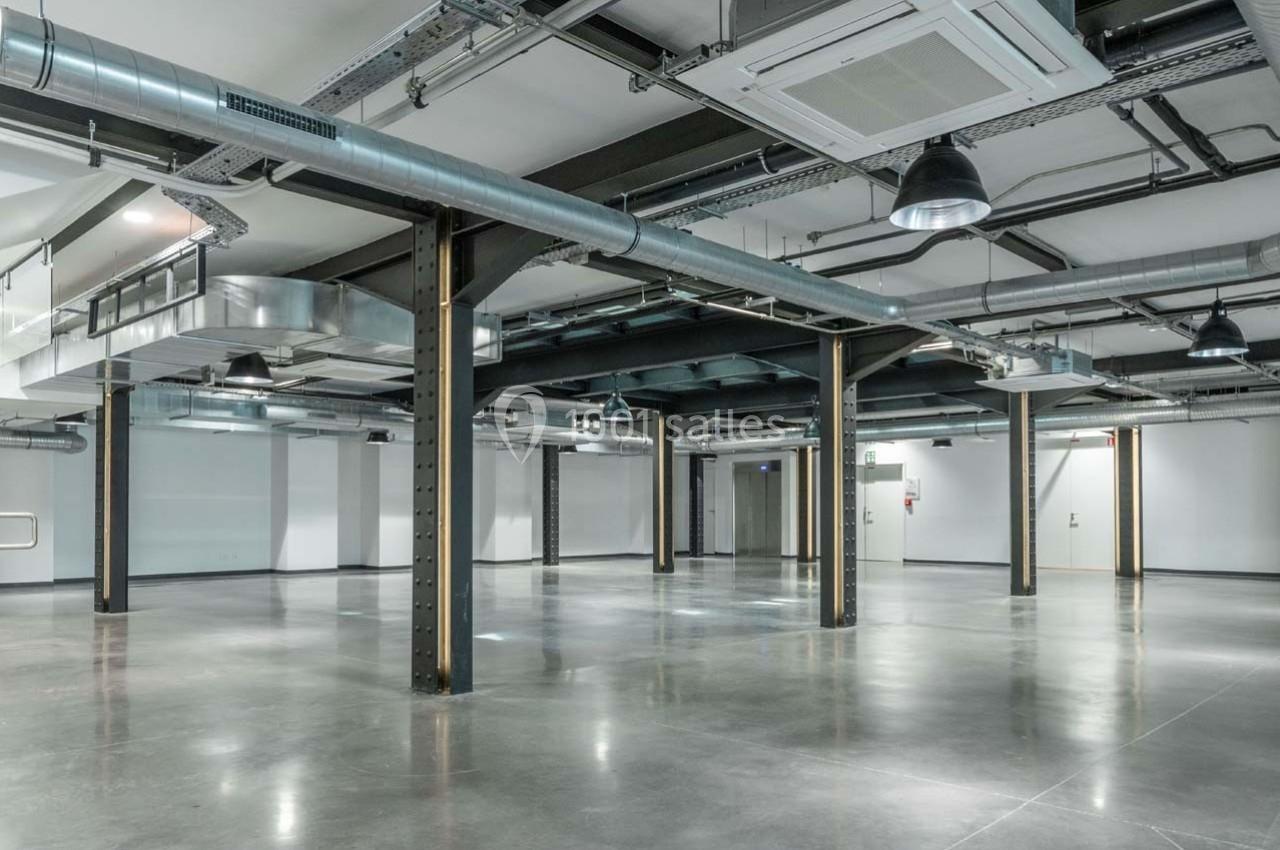 Intérieur d'un espace industriel vide avec sol en béton, poutres métalliques, conduits apparents et éclairage suspendu.