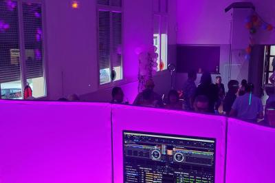Poste de DJ avec une table de mixage, un ordinateur portable affichant un logiciel audio et un micro, éclairé en violet.