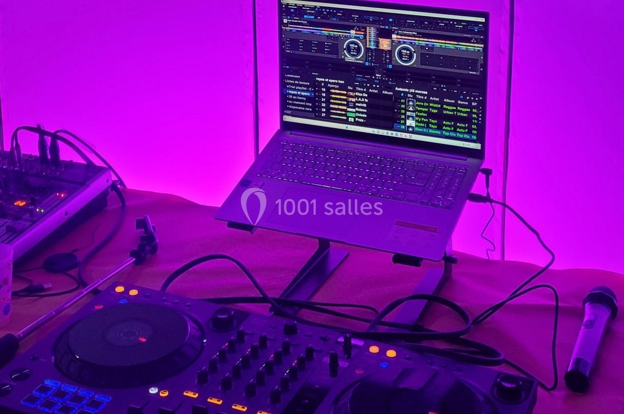 Poste de DJ avec une table de mixage, un ordinateur portable affichant un logiciel audio et un micro, éclairé en violet.