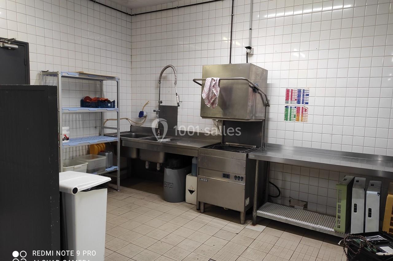 Cuisine professionnelle avec évier inox, étagères de rangement, carrelage mural blanc et sol carrelé beige.