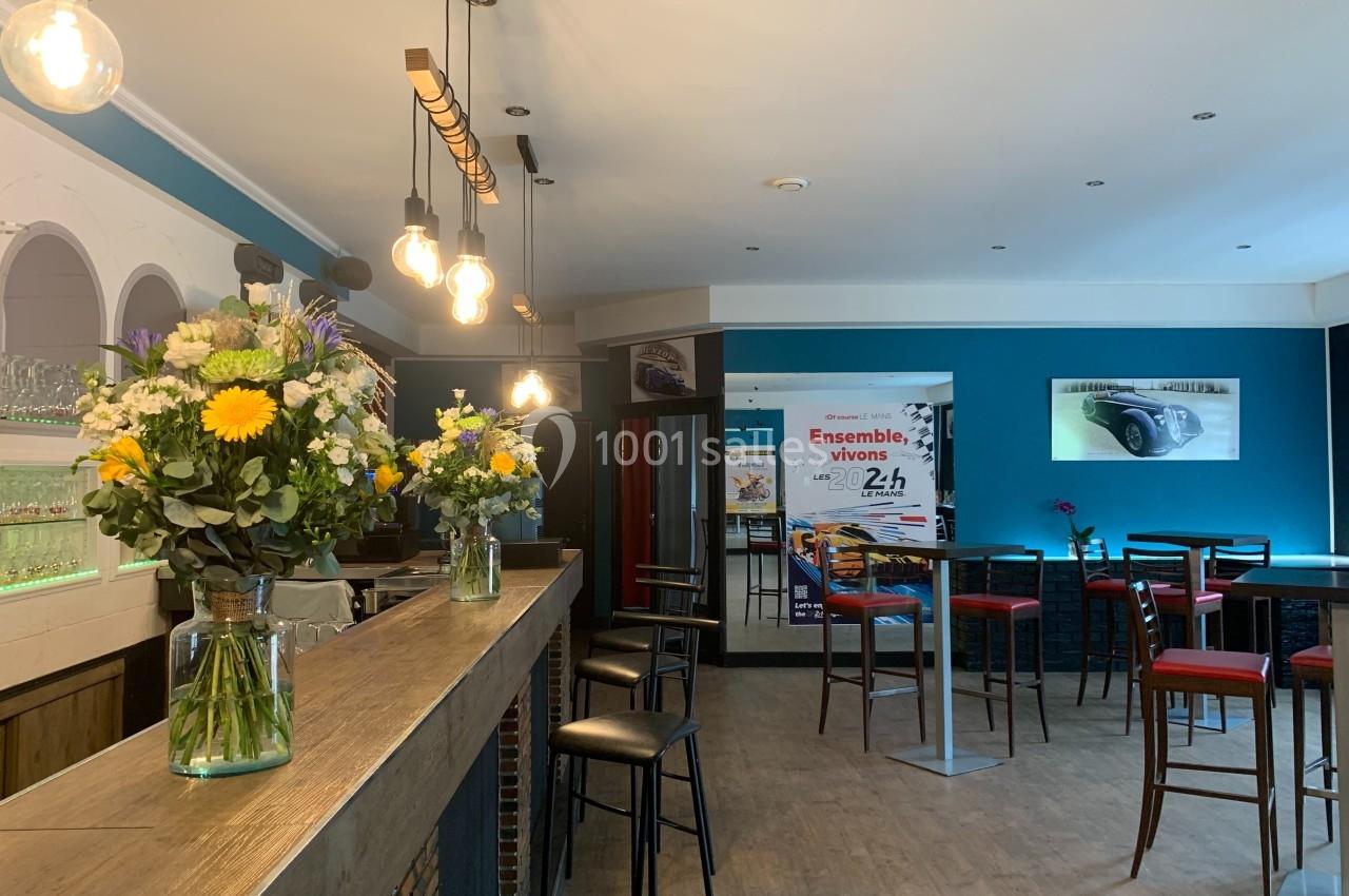 Intérieur d'un café avec un bar en bois, des tabourets, des tables hautes et des bouquets de fleurs colorées.
