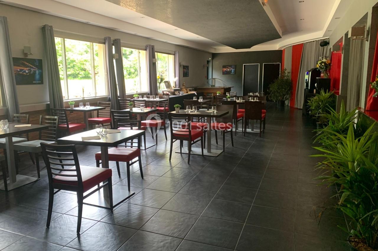 Salle de restaurant lumineuse avec tables dressées, chaises rouges, grandes fenêtres et plantes décoratives.