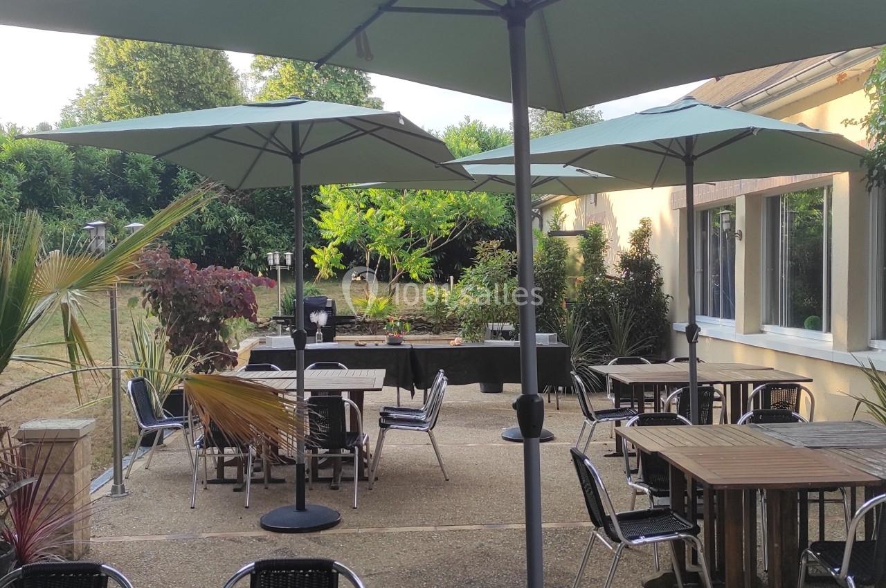 Terrasse extérieure avec tables, chaises et parasols, entourée de verdure et attenante à un bâtiment clair.