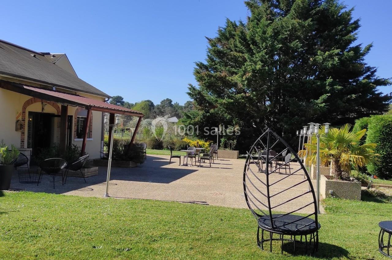 Terrasse ensoleillée avec mobilier de jardin, vue sur un espace vert arboré et une maison en arrière-plan.