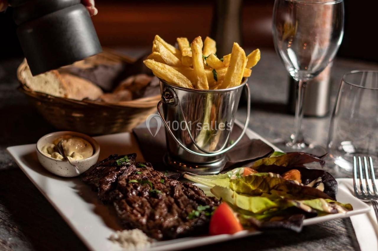 Assiette avec steak grillé, frites dans un seau en métal, salade verte, pain et sauce, sur une table dressée.