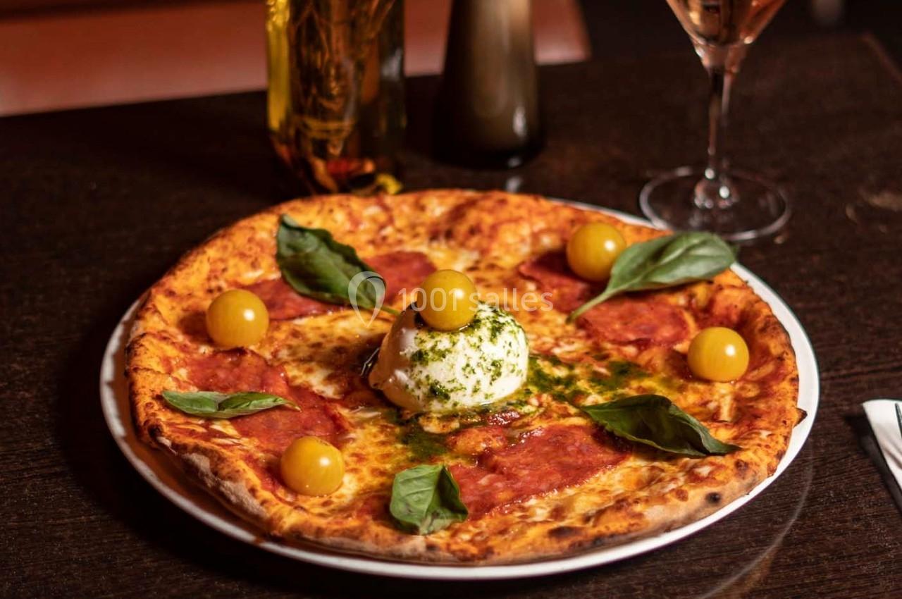 Pizza garnie de mozzarella, tranches de salami, tomates jaunes, feuilles de basilic et sauce pesto, sur une table sombre.