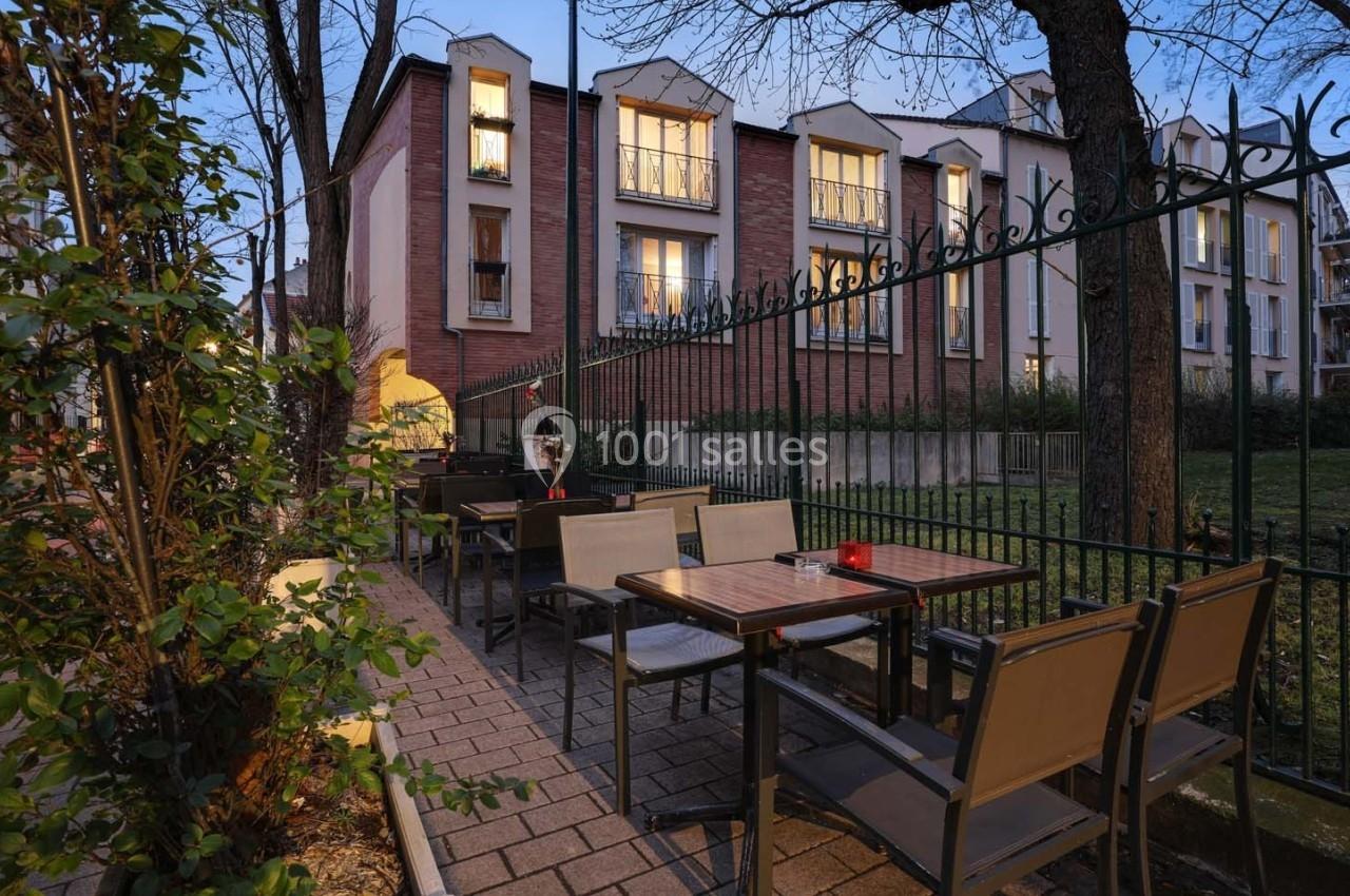 Terrasse extérieure avec tables et chaises, entourée de verdure et clôturée, devant un bâtiment en briques.
