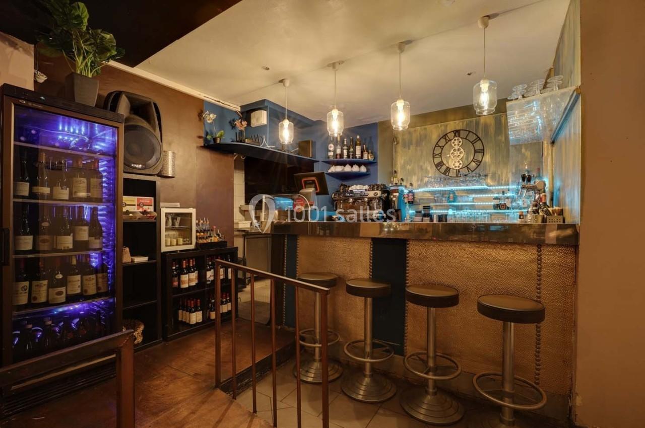 Bar avec comptoir métallique, tabourets hauts, étagères de bouteilles, éclairage suspendu et décoration murale avec horloge.