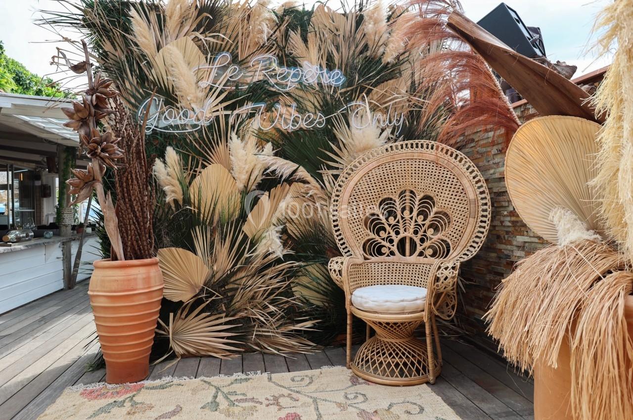 Fauteuil en rotin entouré de plantes séchées et d'une enseigne lumineuse sur une terrasse en bois.