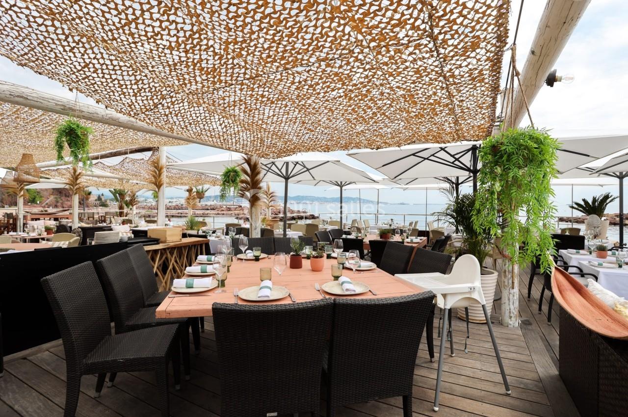 Terrasse d'un restaurant en bord de mer avec tables dressées, chaises en osier, parasols et vue sur l'eau.