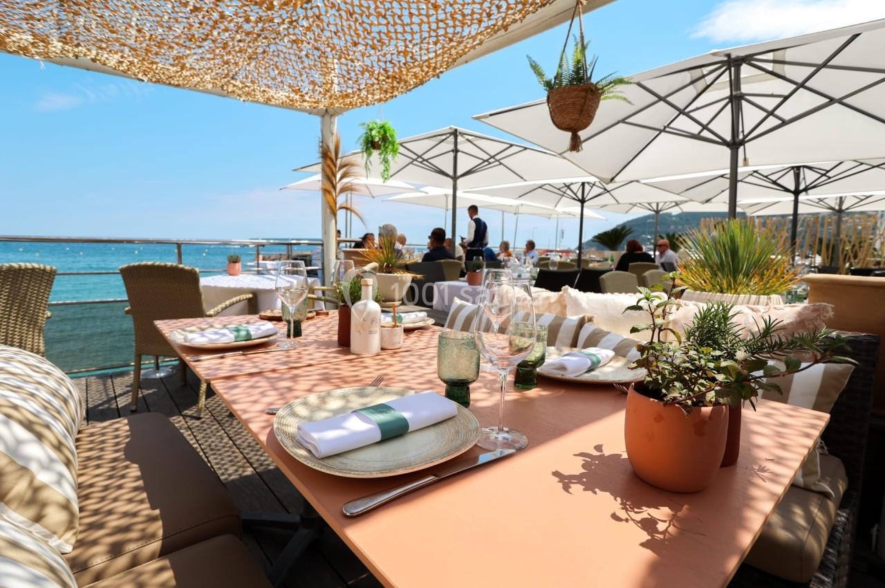 Terrasse d'un restaurant en bord de mer, avec tables dressées, parasols et vue sur l'eau sous un ciel ensoleillé.