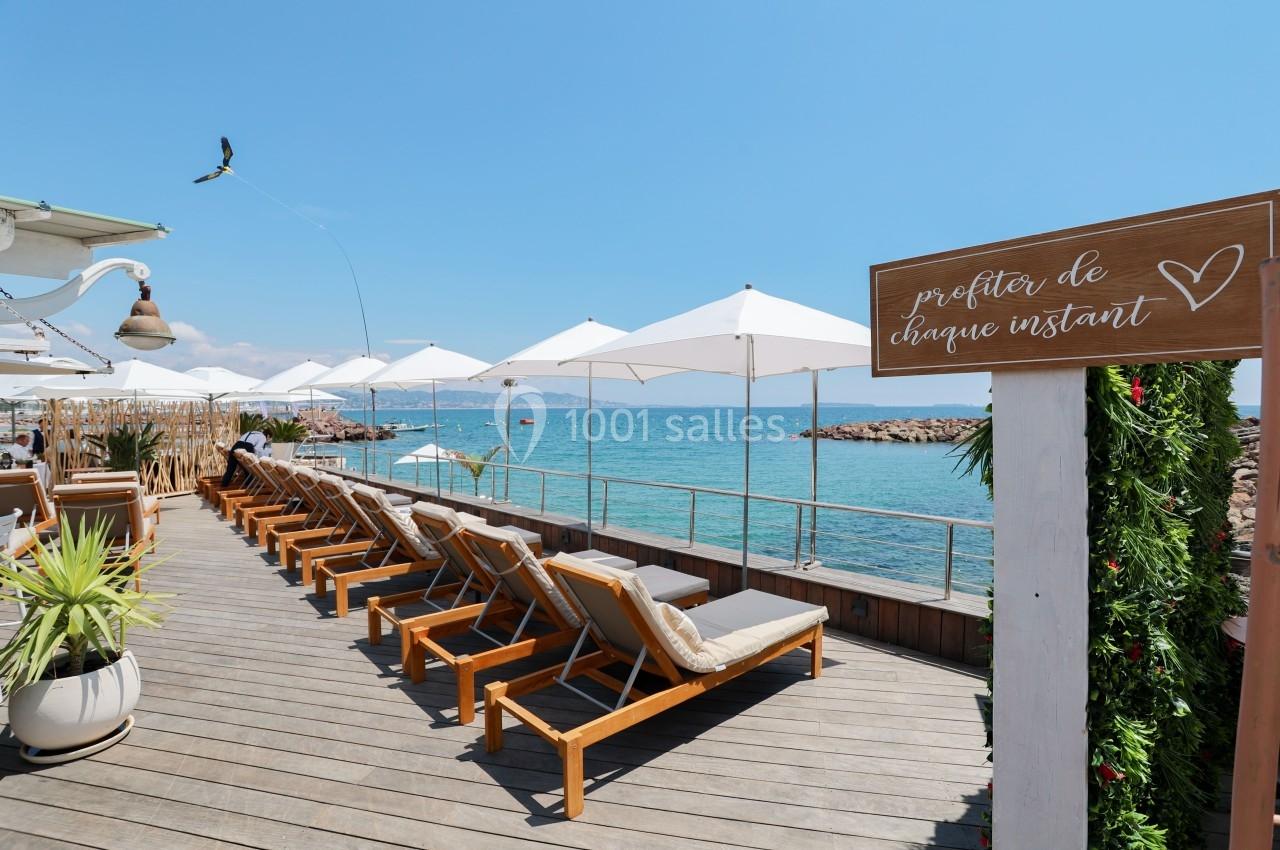 Terrasse en bois avec transats alignés, parasols blancs et vue sur la mer par temps ensoleillé.