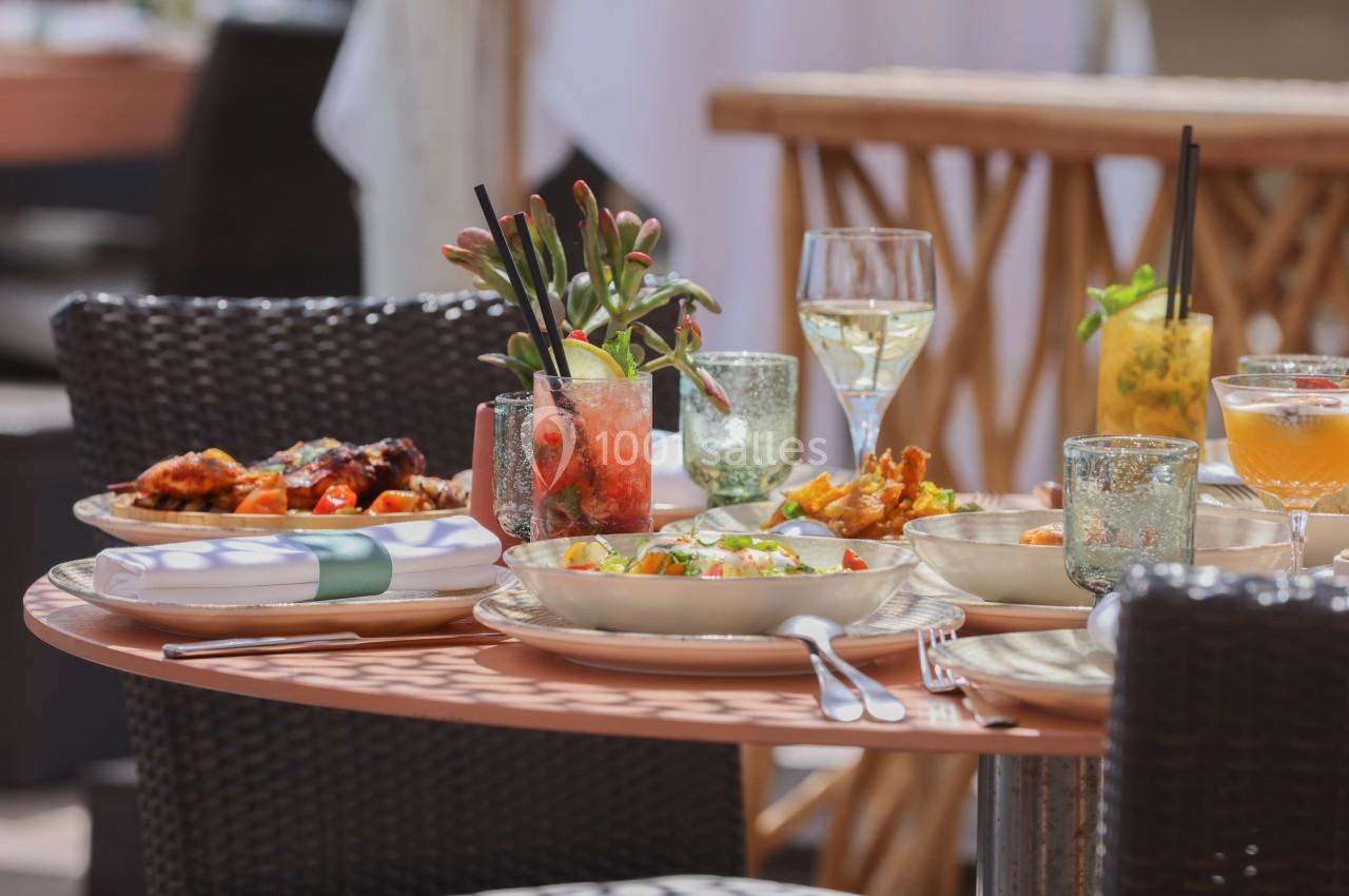 Table en terrasse avec plats variés, cocktails colorés et verres de vin, entourée de chaises en rotin.