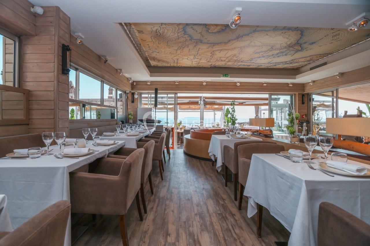 Salle de restaurant lumineuse avec tables dressées, chaises en velours marron et vue sur une terrasse extérieure.