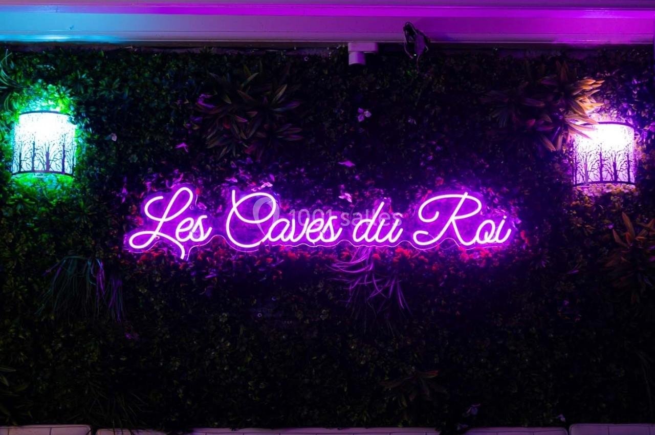 Enseigne néon rose ’Les Caves du Roi’ sur un mur végétal éclairé par deux appliques lumineuses.