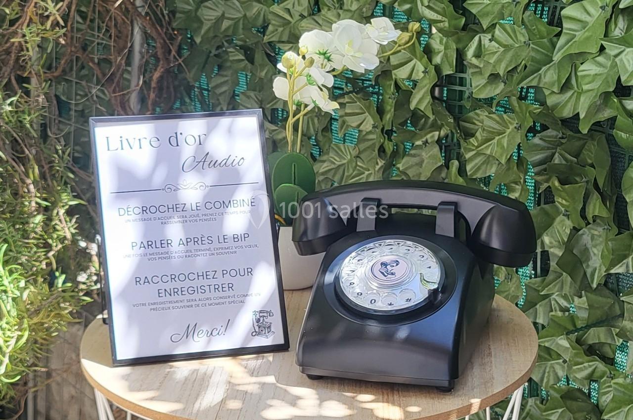 Téléphone rétro noir sur une table ronde en bois, accompagné d'un panneau explicatif et décoré de plantes vertes.
