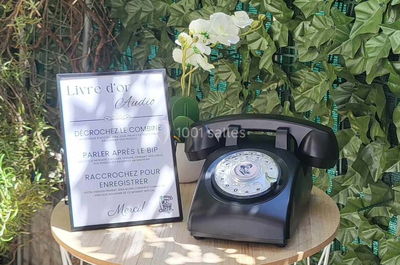 Téléphone vintage noir sur une table ronde en bois, accompagné d'un panneau explicatif et décoré de plantes vertes.