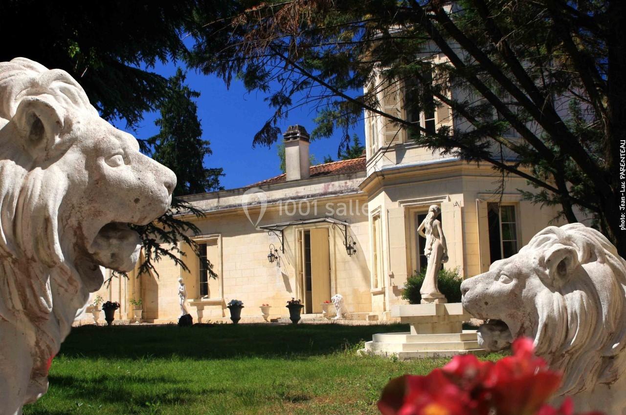 Deux statues de lions en pierre encadrent un manoir lumineux entouré de verdure par une journée ensoleillée.