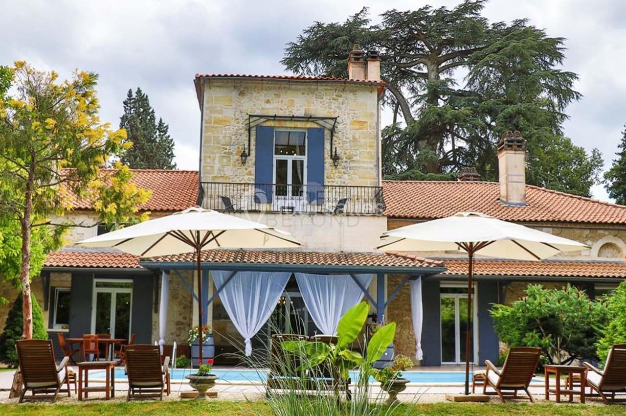 Façade d'une maison en pierre avec terrasse, parasols, chaises longues et piscine, entourée de végétation.