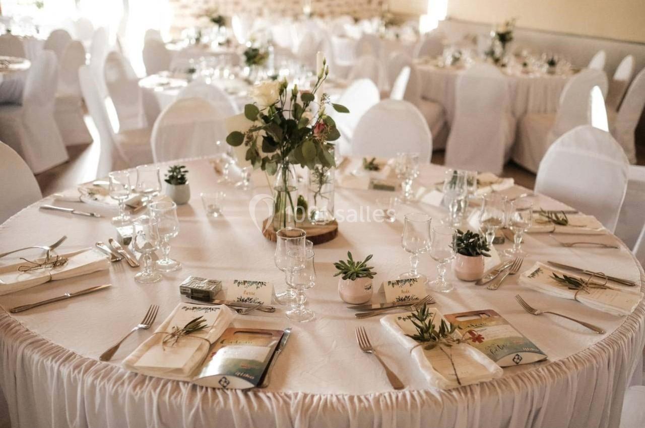 Table ronde décorée pour un événement, avec nappes blanches, vaisselle élégante, menus et centre de table floral.