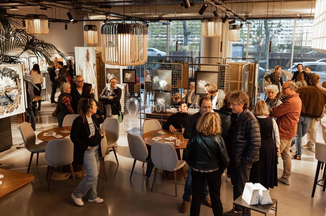 Des personnes discutent et observent des œuvres d'art dans une galerie lumineuse avec des tables et des éclairages suspendus.