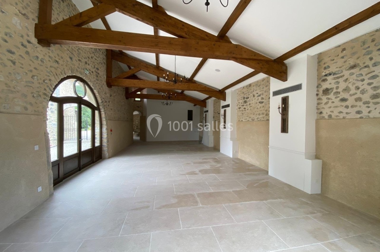 Salle lumineuse avec poutres apparentes, murs en pierre et grandes ouvertures vitrées donnant sur l'extérieur.